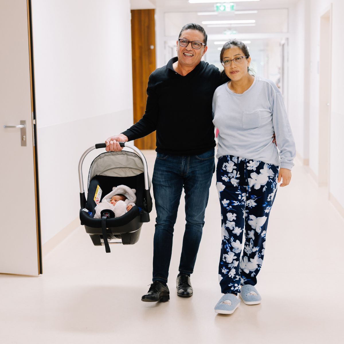 Eltern gehen mit ihrem Neugeborenen im Maxi Cosi entlang des Krankenhausflurs.