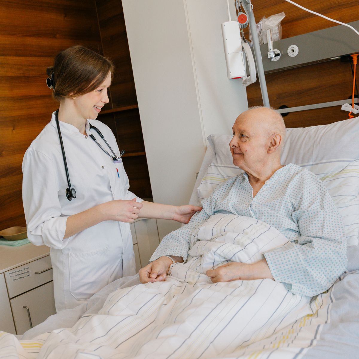 Eine Ärztin steht neben dem Bett eines älteren Patienten. Sie legt ihm die Hand auf den Arm und lächelt.