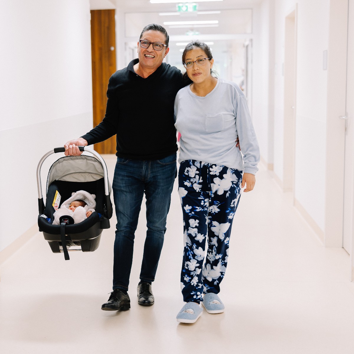 Eltern gehen mit ihrem Neugeborenen im Maxi Cosi entlang des Krankenhausflurs.