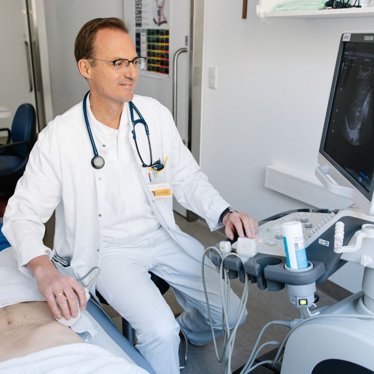 Ein Arzt sitzt an einem Ultraschallgerät und schallt den Bauch eines Patienten, der auf einer Liege liegt.