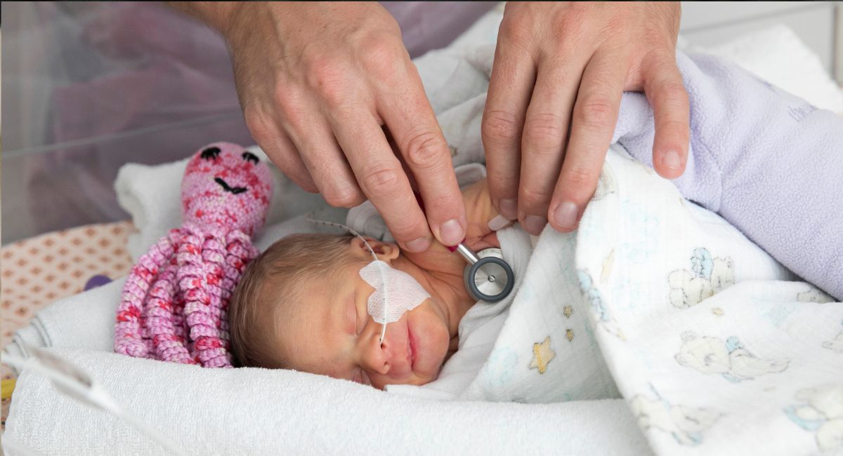 Ein schlafendes Neugeborenes, das auf der Neonatologie untersucht wird. Es trägt ein Herz-Pflaster auf der Wange.