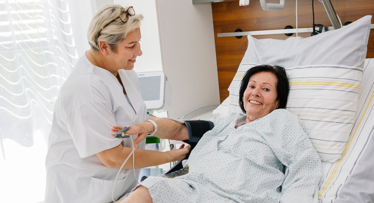 Eine weibliche Pflegekraft misst bei einer Patientin im Bett den Blutdruck.