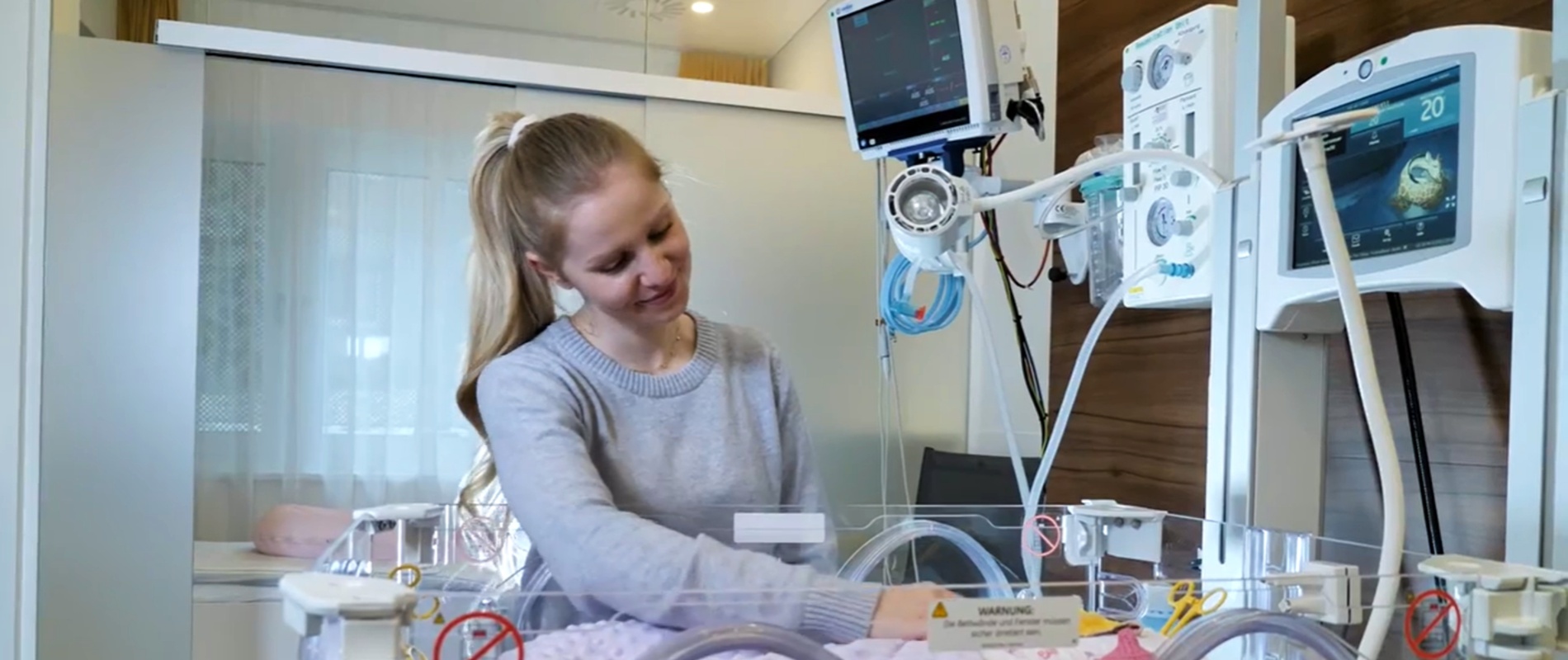 Eine Frau steht vor einem neonatologischen Intensivbett und lächelt das Kind an.