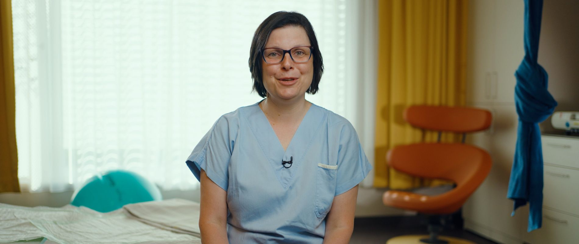 Eine Hebamme sitzt vor einem Krankenbett und spricht in die Kamera.