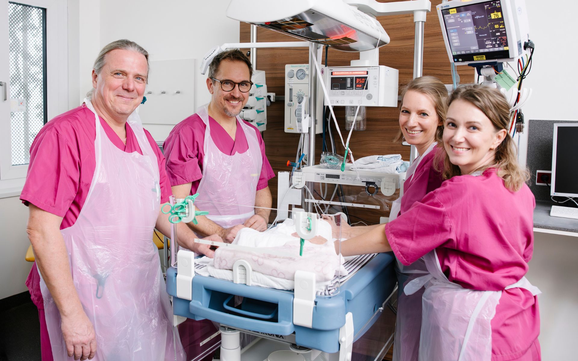 Vier lächelnde medizinische Fachkräfte stehen rund um eine neonatologische Versorgungseinheit.