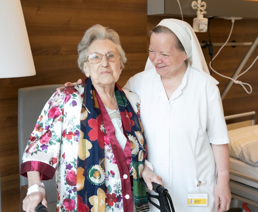 Eine Ordensschwester lächelt eine ältere Patientin an und legt die Hand auf ihre Schulter.