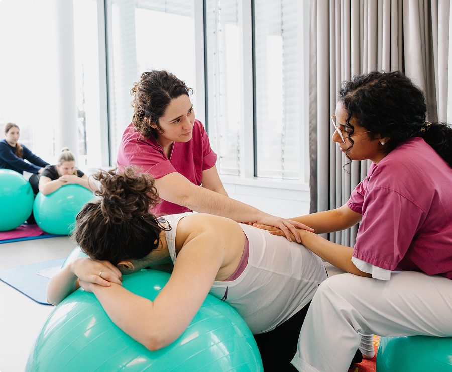 Eine schwangere Frau liegt mit dem Oberkörper auf einem Pezzi-Ball und wird von zwei Physiotherapeutinnen betreut.