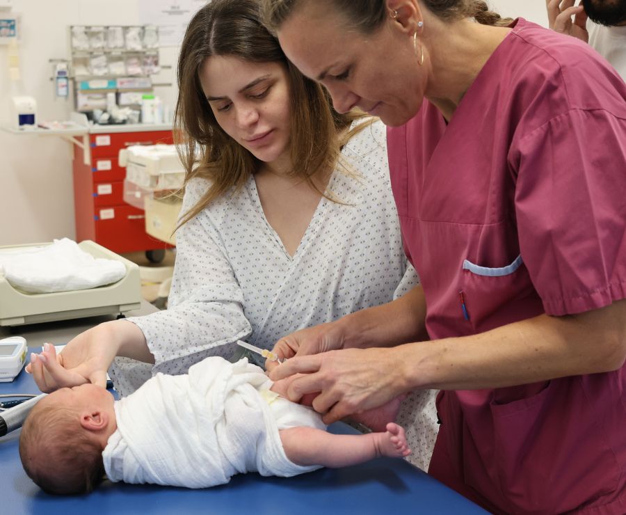 Ein Baby bekommt einen Fersenstich von einer Ärztin. Die Mutter steht daneben. 