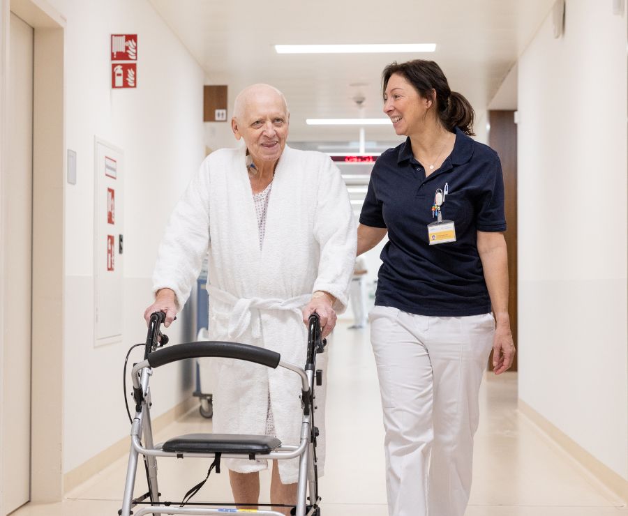 Eine Physiotherapeutin geht mit einem Patienten mit Rollator über den Gang des Krankenhauses.