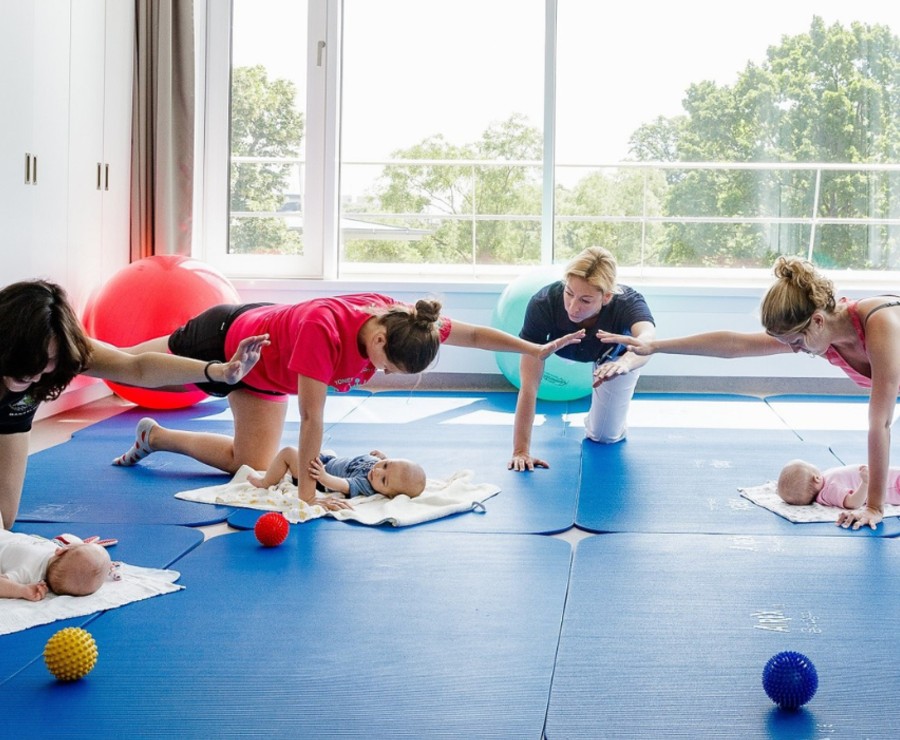 Eine Gruppe Mütter führt auf Matten unter Anleitung ein Training durch, ihre Kinder liegen vor ihnen