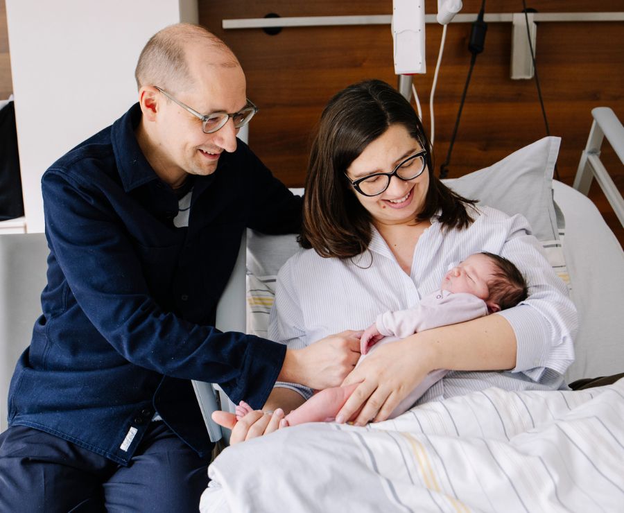 Eine Mutter hält im Bett ihr Neugeborenes im Arm. Der Vater sitzt daneben und streichelt es. Sie lächeln es an.
