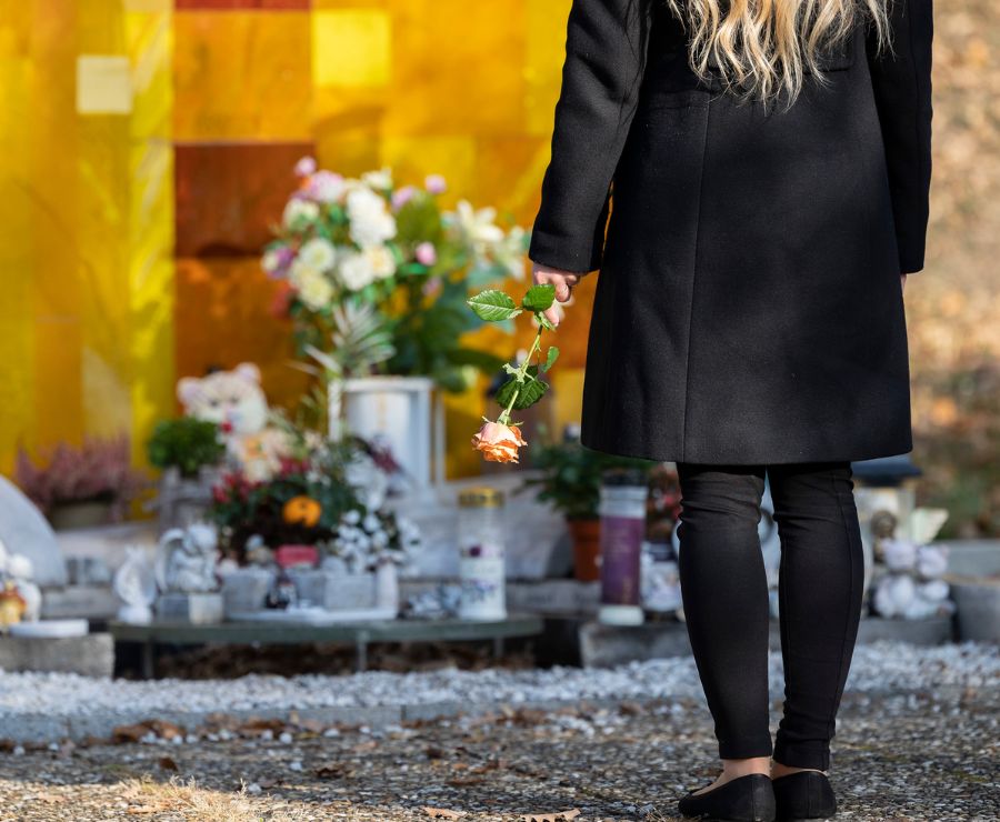 Eine Frau in schwarz steht mit einer Rose in der Hand vor der Grabstätte.