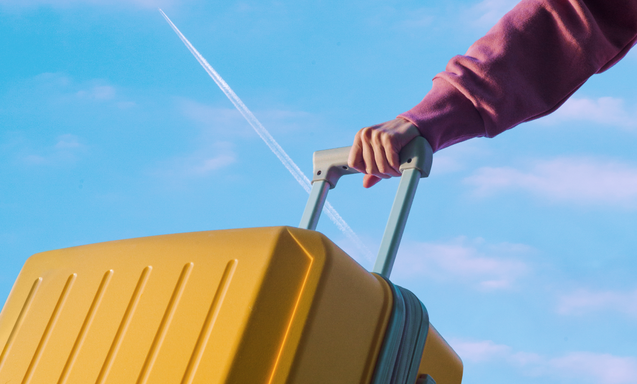 A person in a pink sweater holds a yellow suitcase against a blue sky with a condensation trail from a plane