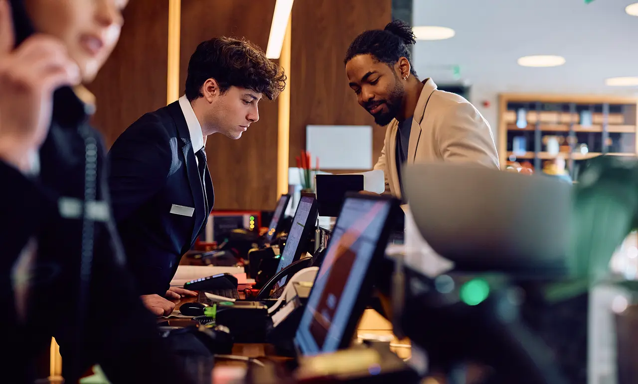Two hotel receptionists in suits assist guests at the front desk, surrounded by computers and phones in a modern lobby setting.