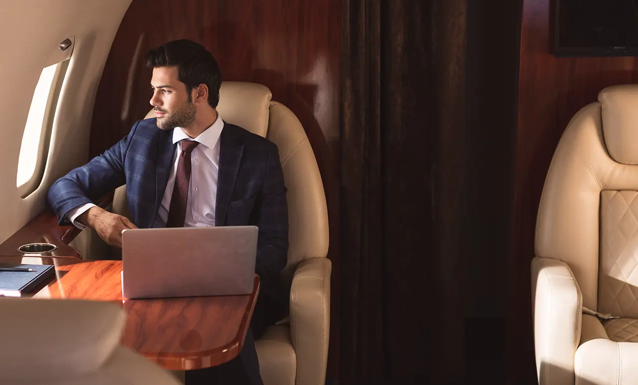 Man in a suit sitting in a private jet, looking out the window, with a laptop on the table beside him.