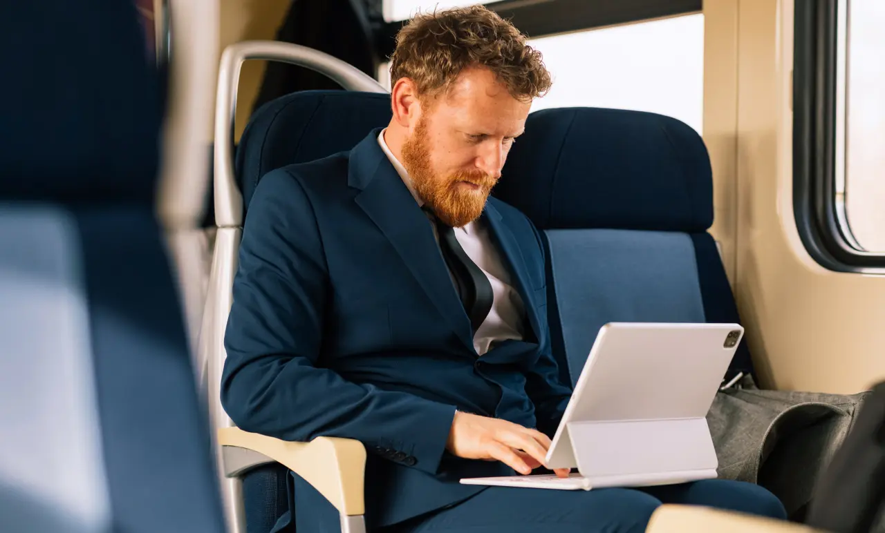 A man in a blue suit works on a laptop while sitting on a train, focused on his screen, with a window view in the background.