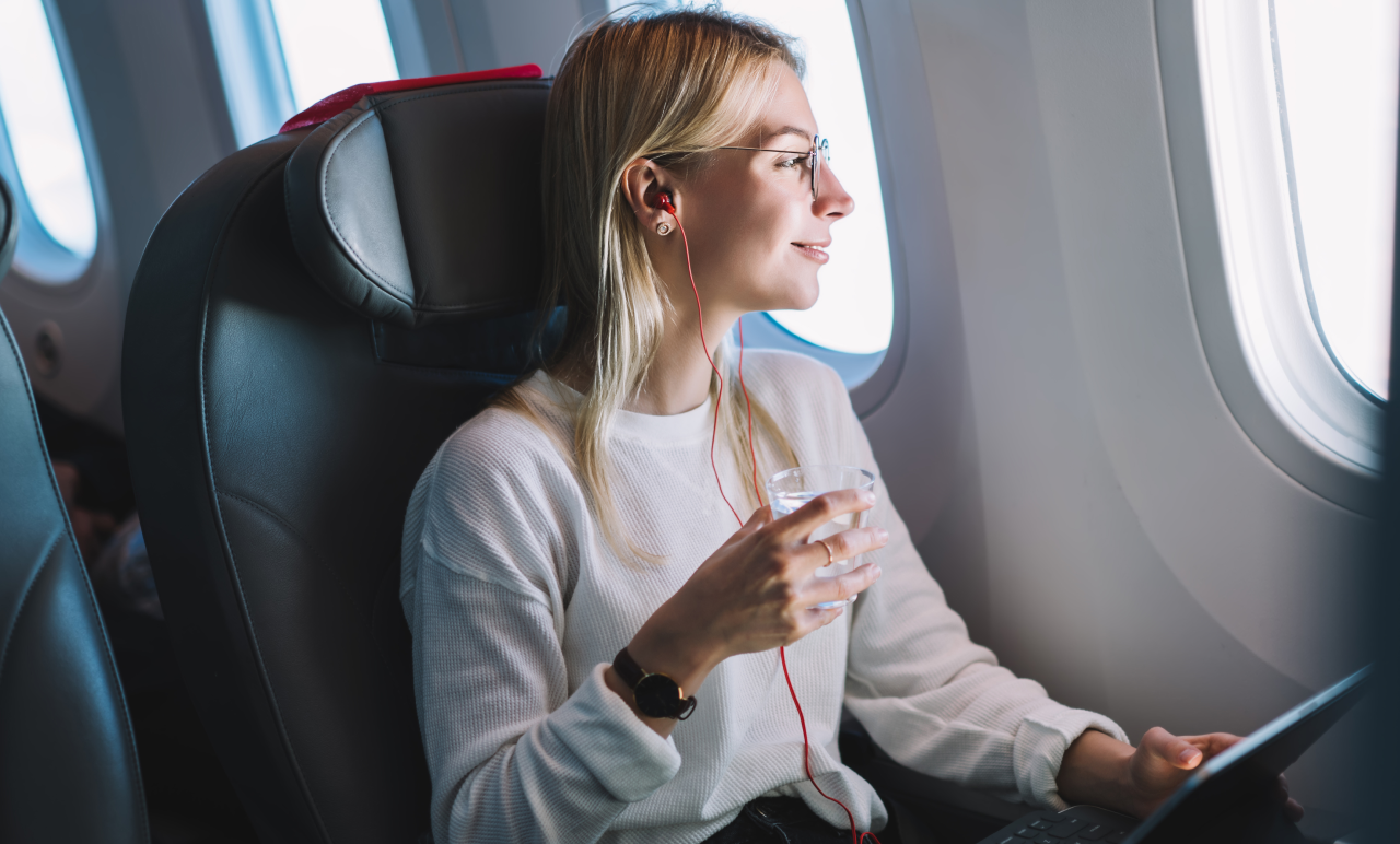 Woman with glasses and earphones sits by airplane window, holding a drink and tablet, looking outside.