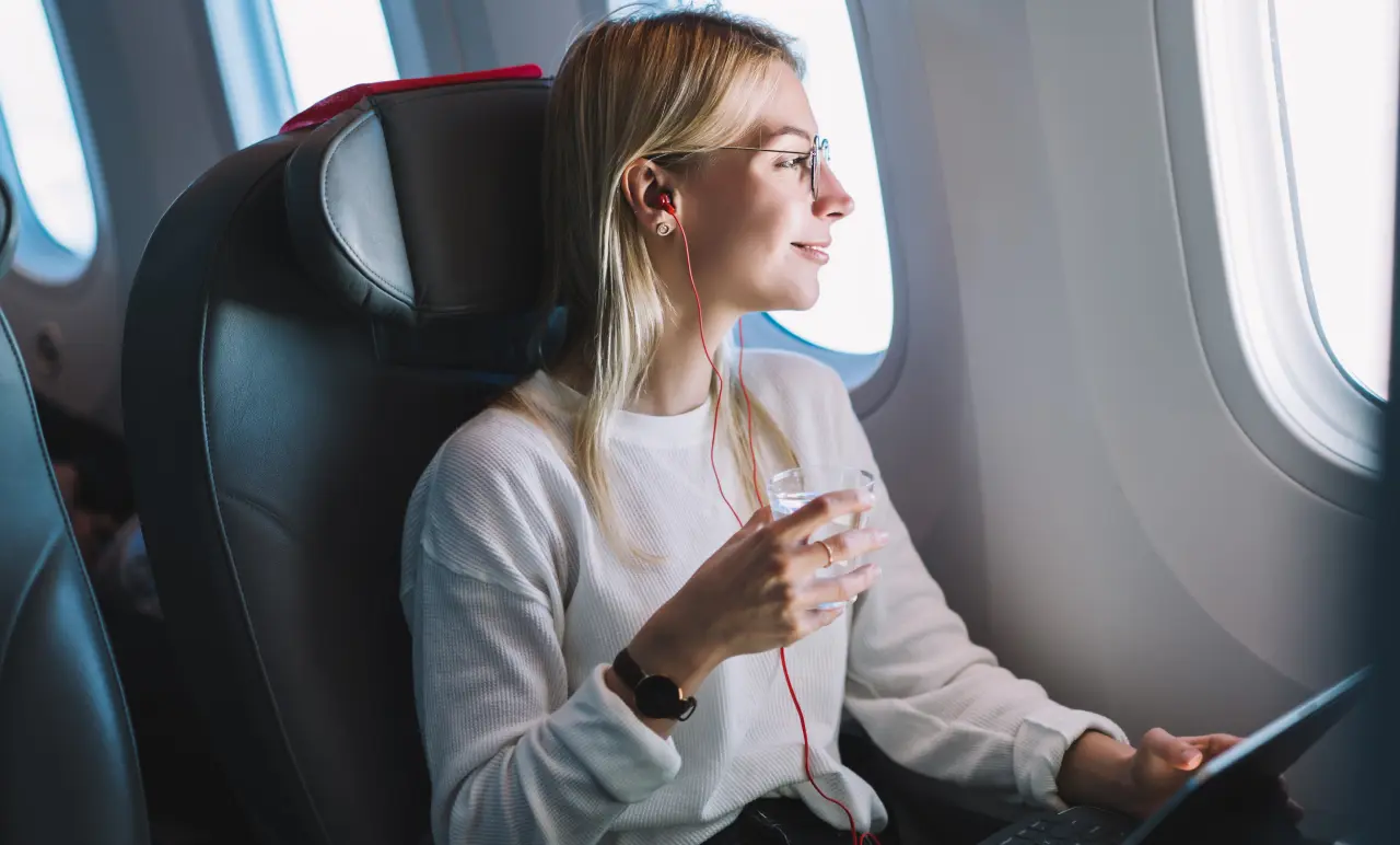 Woman with glasses and earphones sits by airplane window, holding a drink and tablet, looking outside.