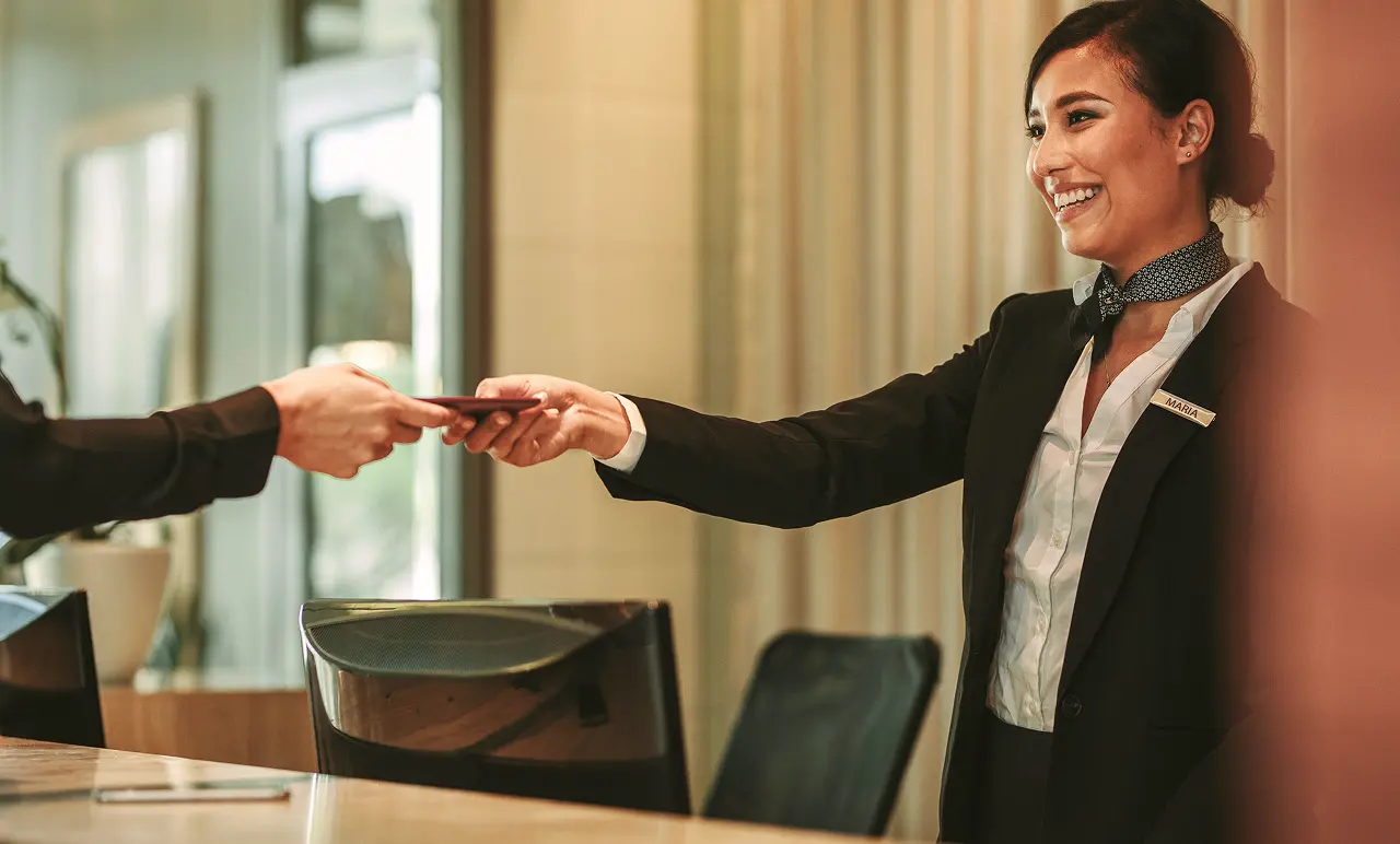 Smiling receptionist in a suit hands a document to a guest across a counter in a modern office setting.