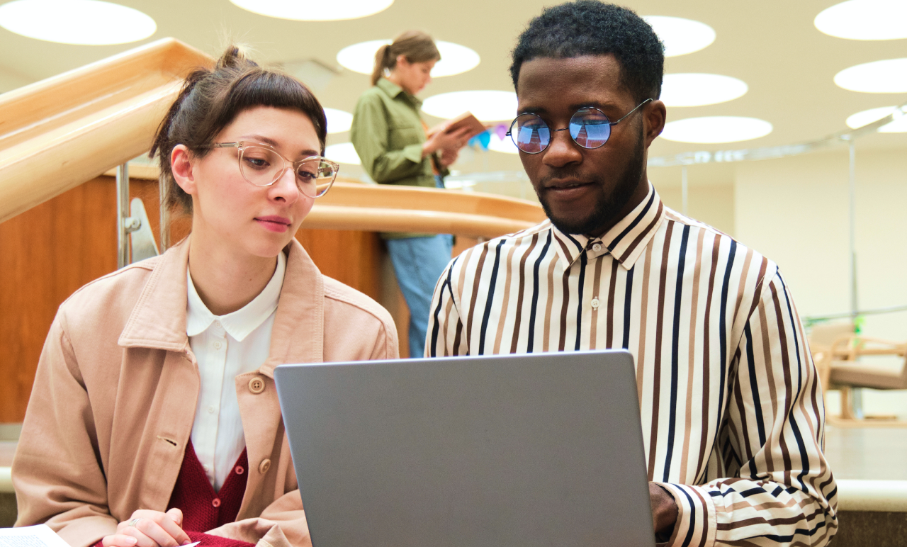Two people sit together, focused on a laptop. The woman wears glasses and a beige jacket, and the man wears glasses and a striped shirt.
