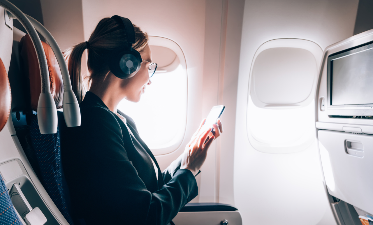 Woman sitting in an airplane seat, wearing headphones, using a smartphone, with sunlight coming through the window.