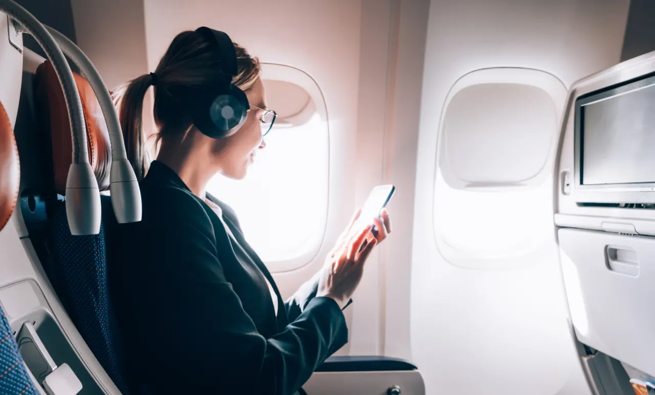 Woman sitting in an airplane seat, wearing headphones, using a smartphone, with sunlight coming through the window.