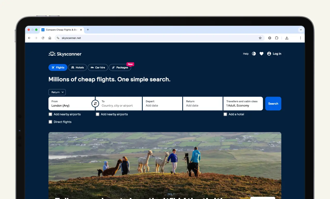 Laptop displaying Skyscanner website with flight search options. Background shows people hiking on a hilltop.