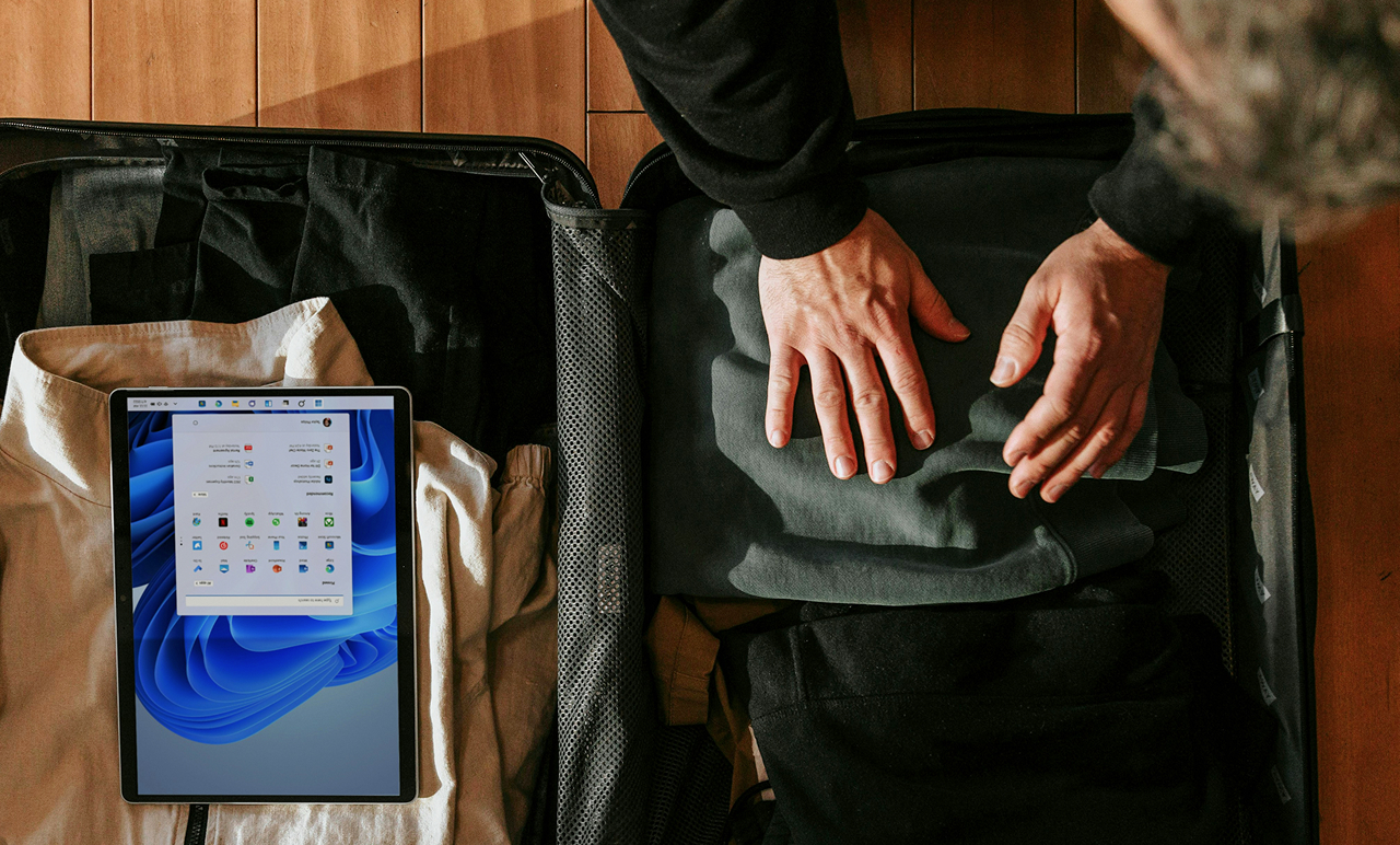 Person packing a suitcase on a wooden floor, with a tablet displaying an operating system screen to the side.