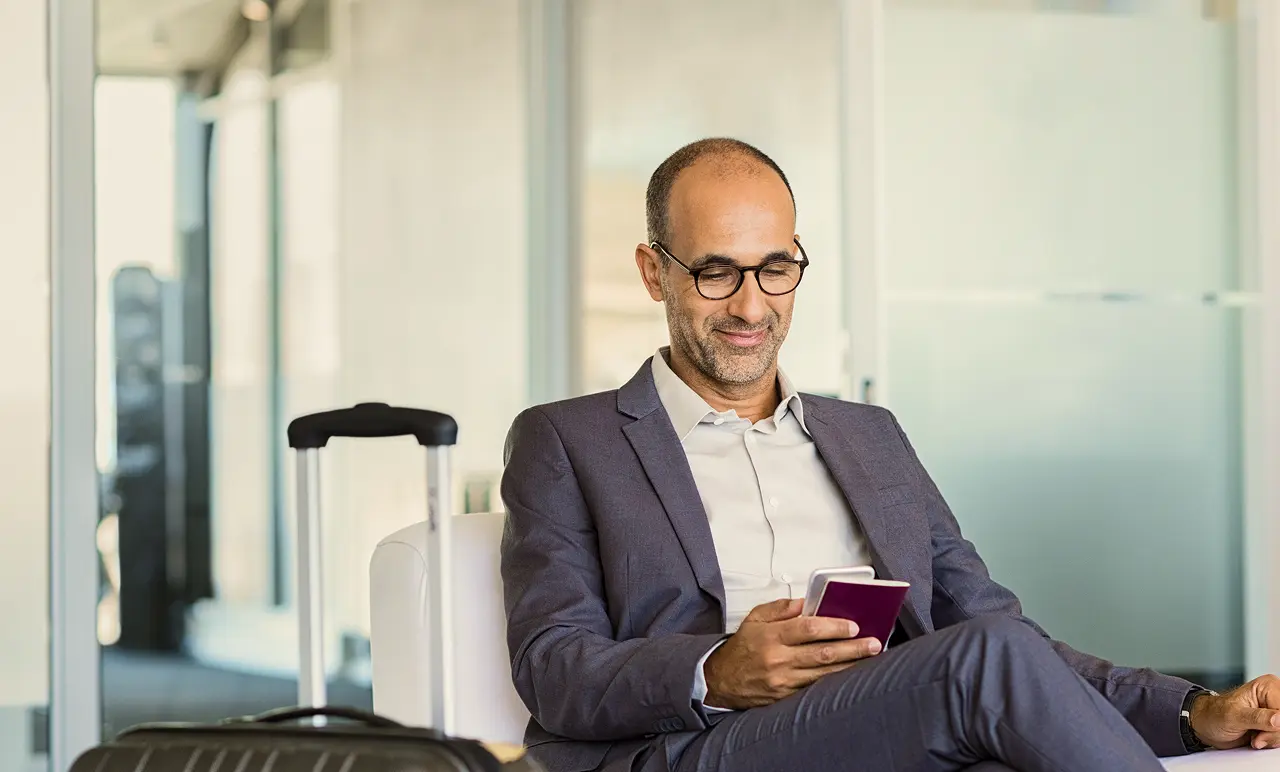 Man in a suit, sitting at an airport lounge, smiling at his phone. A suitcase is beside him. Modern interior background.