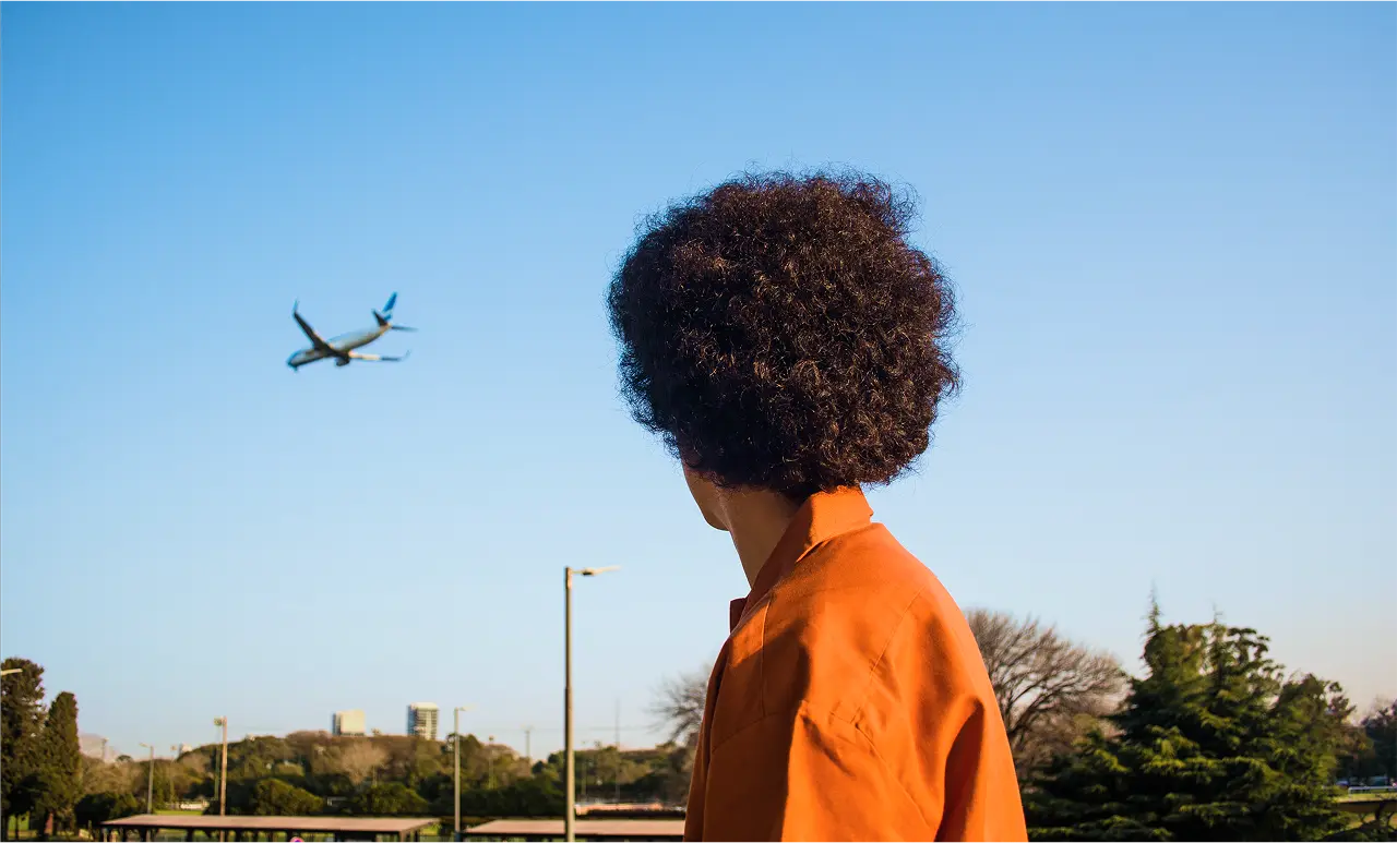Person with curly hair in orange shirt watches airplane flying in clear blue sky, with trees and buildings in the background.