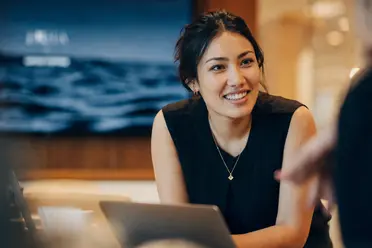 A woman with dark hair wearing a black top smiles while working on a laptop in a modern office setting.
