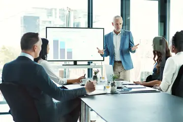A man in a blue blazer presents data on a screen to a group of seated colleagues in a modern conference room.