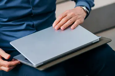 A person in a blue shirt holds a closed laptop with one hand resting on the top, sitting on a chair.