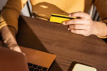 A person in a yellow sweater holds a yellow credit card while using a laptop on a wooden table.