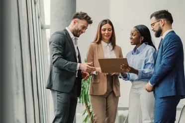 Four business professionals in suits standing and discussing a document together in a modern office setting.