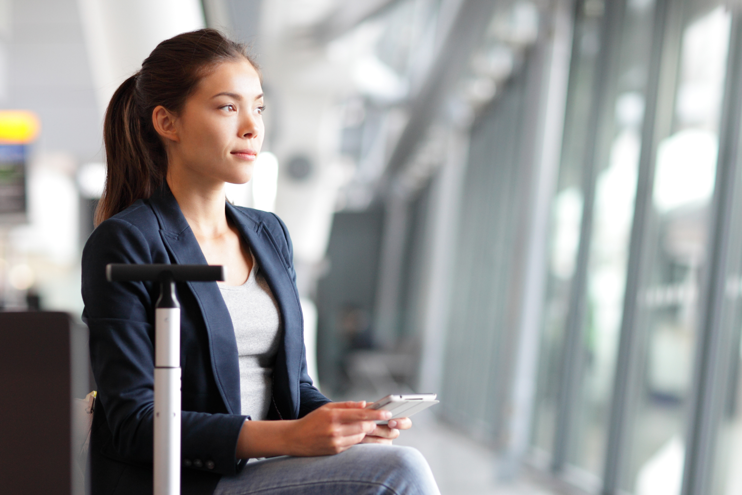 Woman sitting in an airport terminal, holding a tablet, looking out the window, with a suitcase beside her.