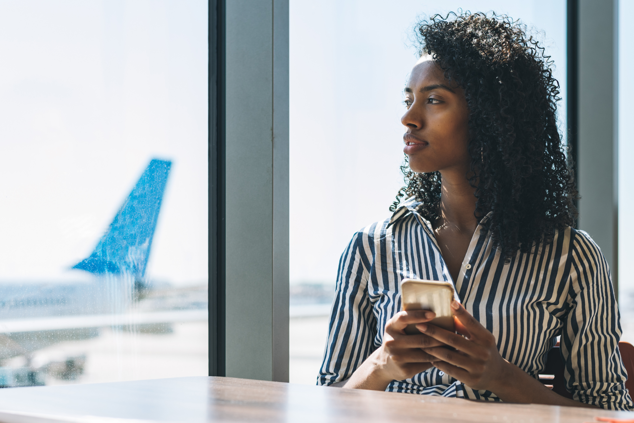Woman with curly hair holding a phone, looking out a window at an airport, with an airplane visible outside.