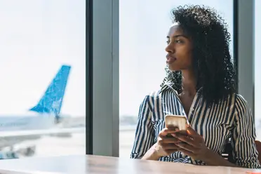 Woman in a striped shirt holding a phone, gazes out an airport window. An airplane is visible outside on the tarmac.