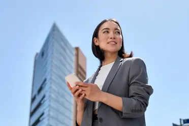 A woman in a gray suit holding a smartphone, standing confidently against a clear blue sky with a modern building in the background.