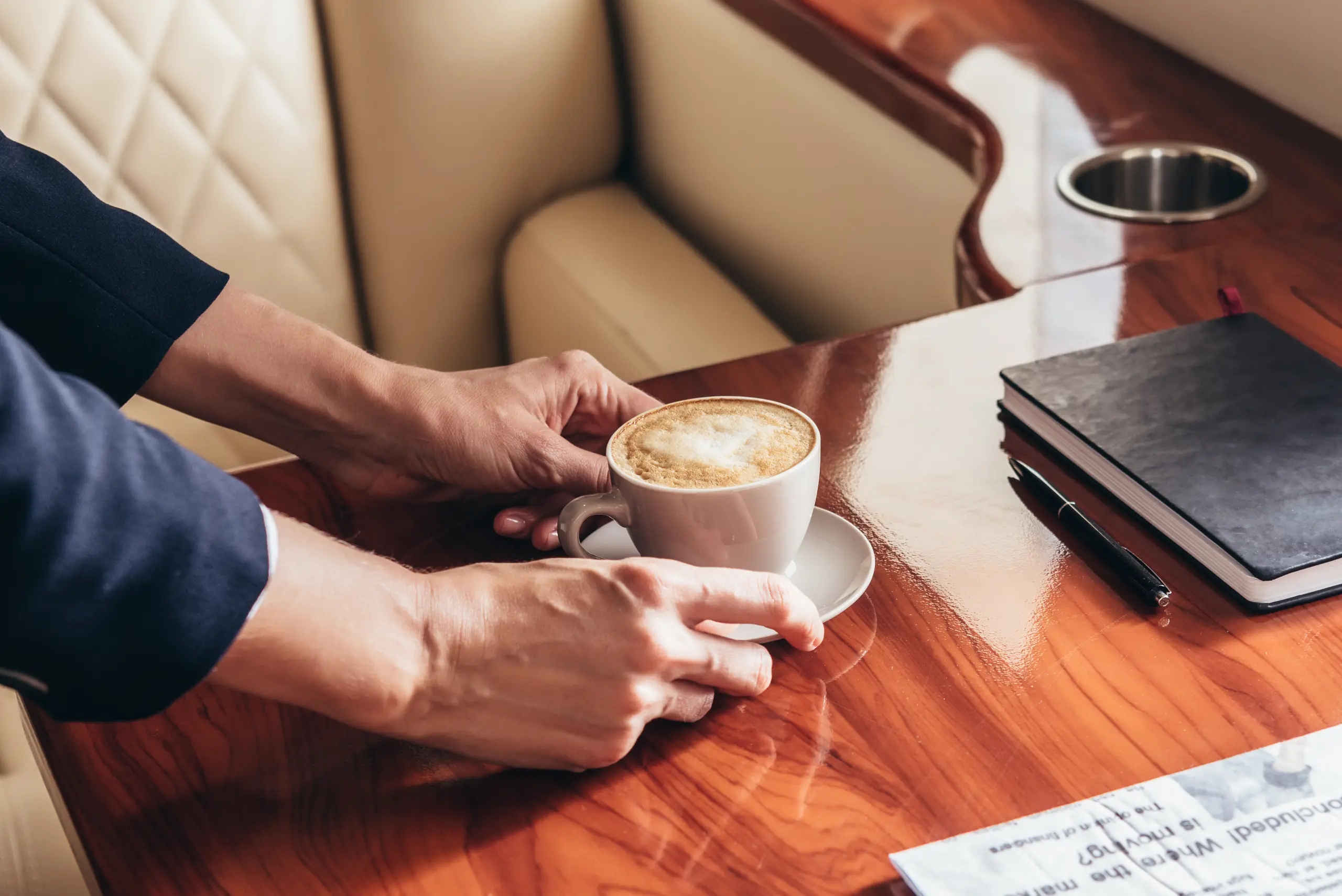 Hands placing a cup of cappuccino on a wooden table over a private jet seat, with a notebook and pen nearby.
