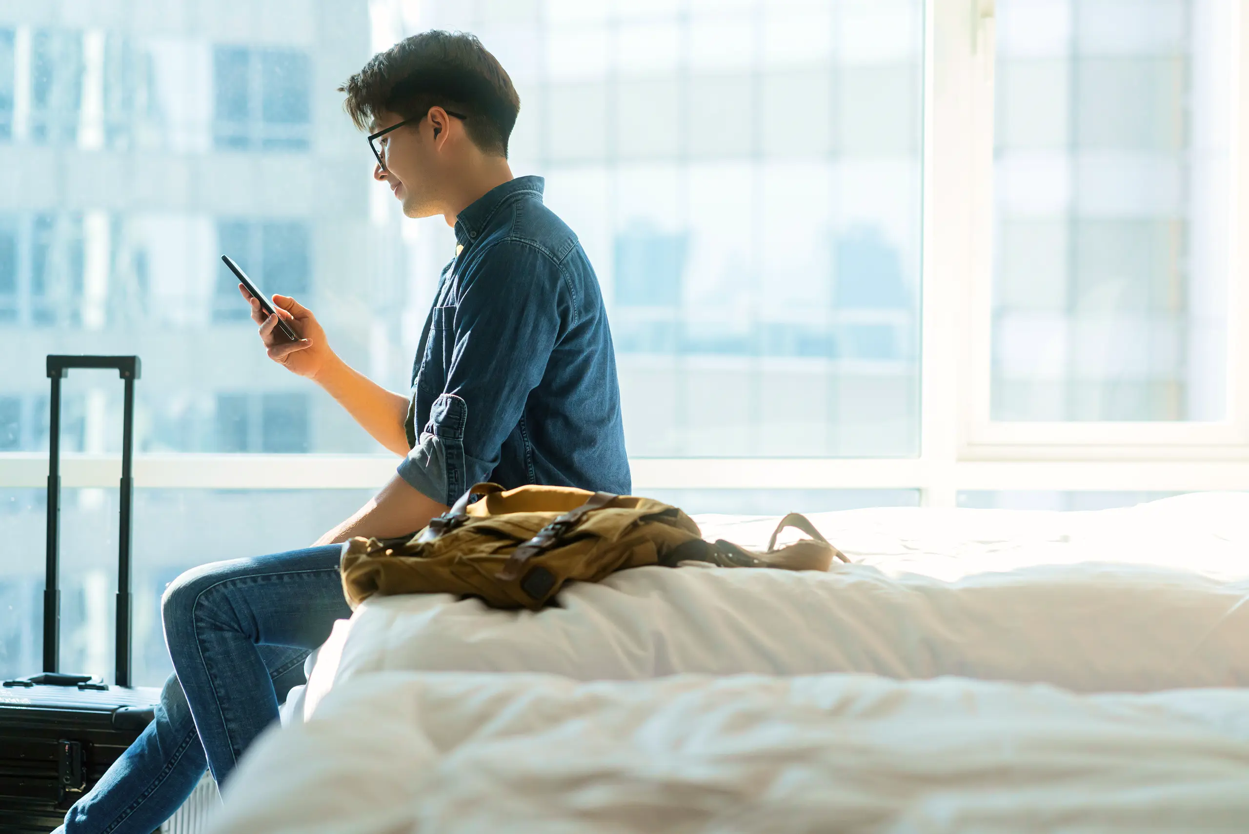 Young man in denim, sitting on a bed in a bright room, checks his phone. A suitcase and backpack are nearby, with cityscape visible outside.