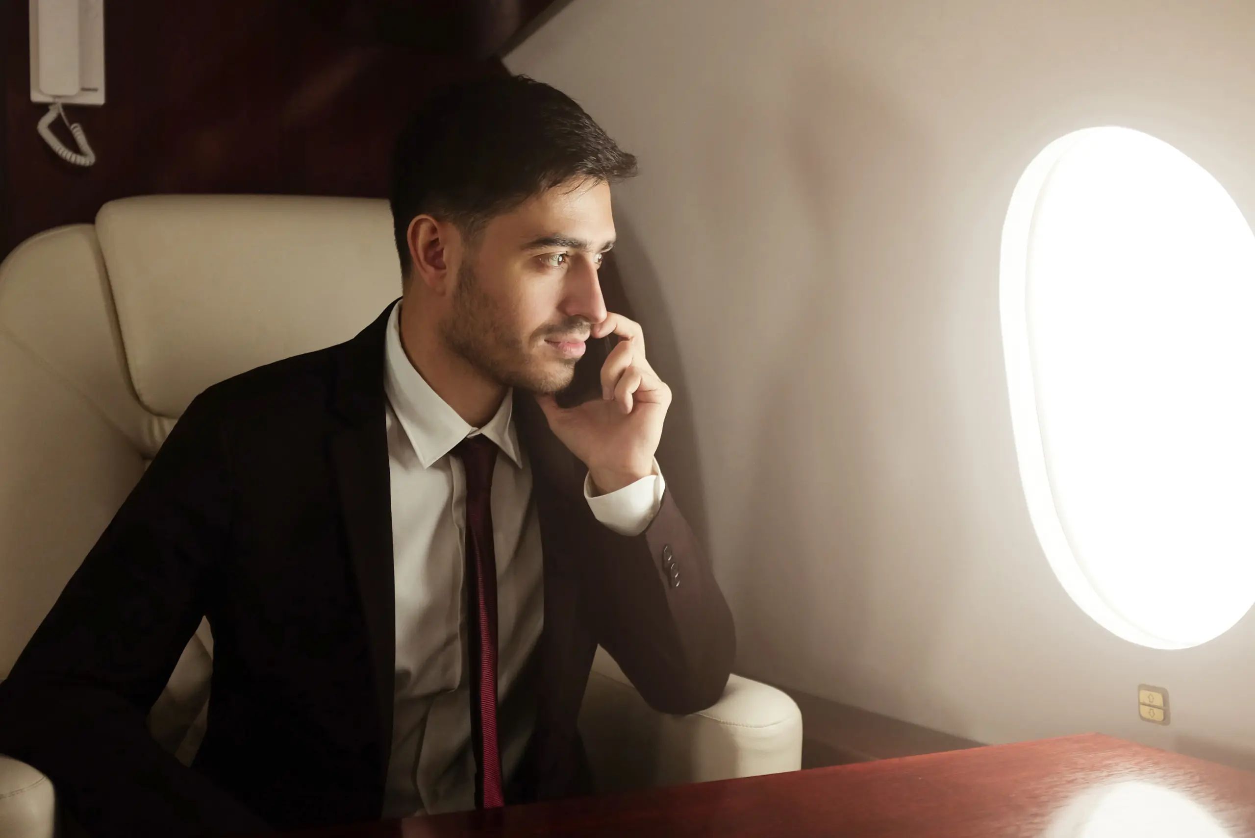 Man in a suit talking on a phone while seated in a private jet, looking out of the window at bright light.