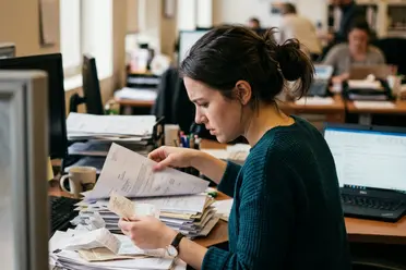 Woman in a green sweater examines papers at a cluttered desk in a busy office, with computers and files around her.