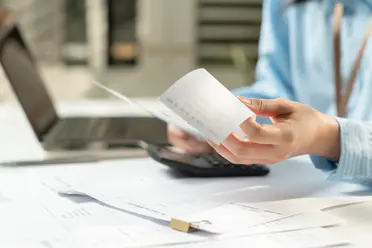 Person holding receipts and using a calculator, with papers and a laptop on a desk, suggesting financial or office work.