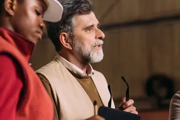 Two people in a conversation, one holding glasses, wearing casual vests inside an industrial setting.