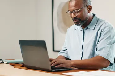 A man with glasses and a beard is focused on typing on a laptop at a desk, with a modern abstract artwork in the background.