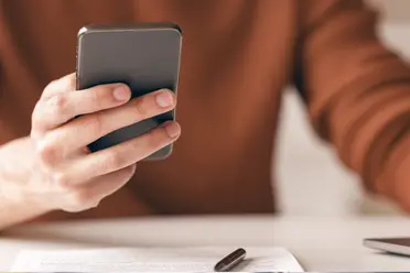 A person in a brown sweater holding a smartphone, with a pen and document on a white desk.