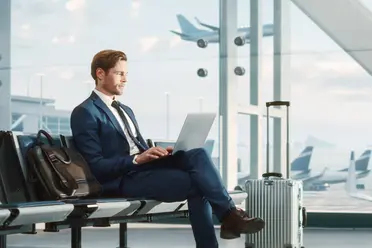 Man in a blue suit sits in an airport lounge using a laptop, with a suitcase beside him and planes visible through large windows.