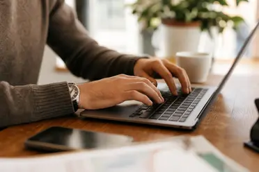 Person typing on a laptop at a wooden table, with a smartphone, papers, and a coffee cup nearby.