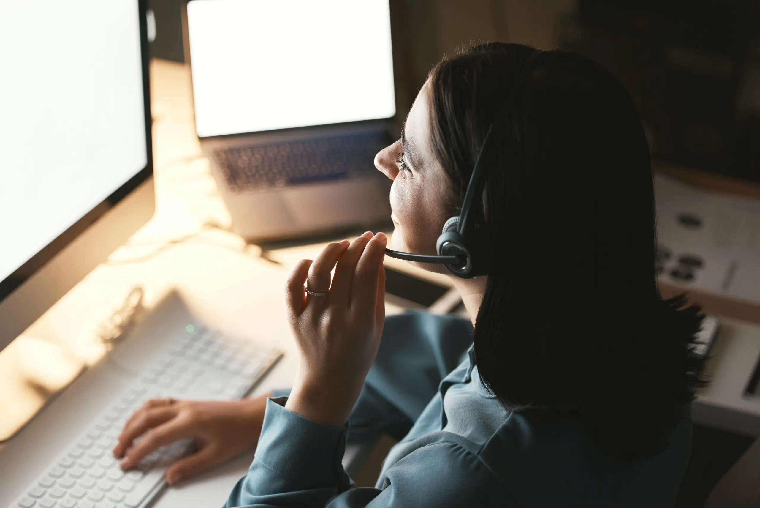 Woman with a headset working on a computer, illuminated by screen light, with a laptop in the background.